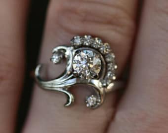 Könnte beinhalten: Ein silberner Ring mit einem großen, runden, klaren Diamanten. Der Ring hat ein dekoratives, wirbelndes Design mit kleineren Akzentsteinen. Der Ring befindet sich an einem Finger. Das Design ist im Jugendstil gehalten.