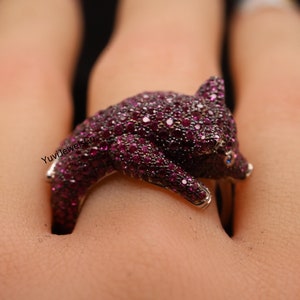 May include: A ring featuring a detailed pig design, crafted from silver and set with sparkling red gemstones. The pig is positioned on its side, with its head turned to the side, and its eyes are accented with blue gemstones.