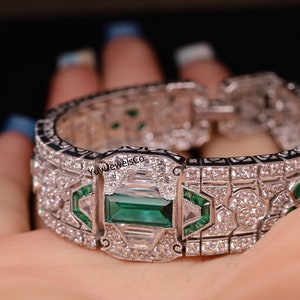 Op de afbeelding: Een zilveren armband met een rechthoekige smaragdgroene edelsteen omringd door kleinere diamanten en smaragdgroene stenen. De armband heeft een gedetailleerd ontwerp met zwarte emaille accenten.