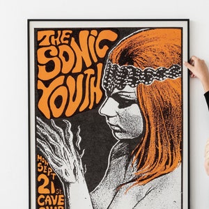 May include: A framed vintage poster with the words "THE SONIC YOUTH" in orange lettering. The poster features a black and white illustration of a woman with orange hair and a decorative headband. The poster also includes event details.