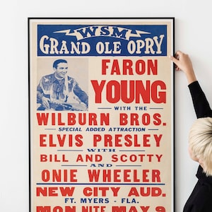 May include: A framed vintage poster for a Grand Ole Opry show featuring Faron Young, Elvis Presley, and others. The poster has a cream background with red and blue text. The poster is being held by a person.