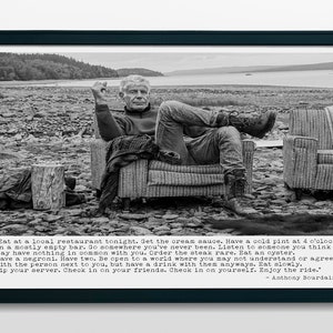 May include: Black and white framed print featuring a seated man in a chair, with a quote by Anthony Bourdain. The image is set outdoors with a rocky landscape and water in the background. The quote is in a typewriter-style font.