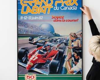 Puede incluir: Un póster vintage enmarcado para la carrera del Gran Premio Labatt du Canada, con un coche de carreras rojo en primer plano. El póster tiene un fondo azul con texto naranja y la fecha "11-12-13 juin 82". Una persona sostiene el póster.
