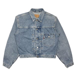 May include: A light blue denim jacket with a button closure, two front pockets, and a single chest pocket. The jacket has a classic style and is made of a durable material.
