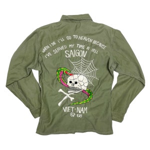 Puede incluir: Una camisa verde de estilo militar con una calavera y una serpiente bordadas en la espalda. El texto "WHEN I DIE I'LL GO TO HEAVEN BECAUSE I'VE SERVED MY TIME IN HELL SAIGON VIET-NAM 67-68" también está bordado en la espalda.