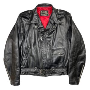 May include: Black leather jacket with a red quilted lining. It has a zip front, multiple pockets, a belt with a buckle, and a collar. The jacket has a classic biker style with silver hardware and the text "The Leather Shop" on the label.