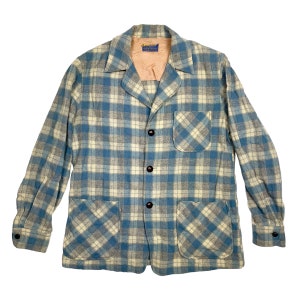 Op de afbeelding: Een blauw en wit geruit jack met een knoopsluiting en twee klepzakken. Het jack heeft een kraag en lange mouwen.
