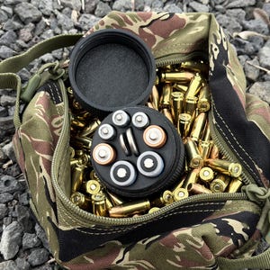 May include: A camouflage-patterned bag filled with brass ammunition and a black container holding various batteries. The bag is open, revealing the contents, and sits on a bed of gray gravel.
