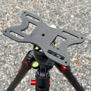 May include: A black metal camera tripod head with a unique, open design. The head features multiple cut-out sections and mounting holes. The tripod has red and green level indicators and red adjustment knobs. The legs are black with red accents.