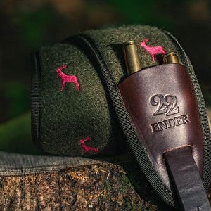 Könnte beinhalten: Ein dunkelgrüner Patronenhalter aus Filz mit braunem Lederakzent. Der Halter hat gestickte rosa Hirsche und den Text "22 ENDER". Zwei Messingpatronen sind sichtbar.