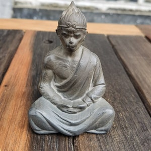 Pode incluir: Uma estatueta cinzenta de uma figura sentada semelhante a um macaco em pose meditativa. A estátua tem um adorno de cabeça detalhado e é coberta por uma túnica. A estatueta é colocada sobre uma superfície de madeira.