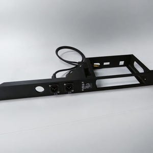 Może przedstawiać: Czarny metalowy element elektroniczny z prostokątną ramą i różnymi portami i złączami. Otwór kołowy i dwa porty wejściowe są widoczne z przodu. Do elementu przymocowany jest czarny kabel.