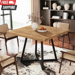 May include: A square dining table with a light brown wood grain top and a black metal base featuring angled legs. The table is surrounded by four wooden chairs with beige cushions. A white vase with dried flowers and a bowl of fruit sit on the table.