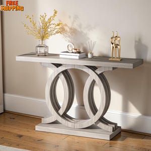 May include: A gray console table with a geometric design. The table features a rectangular top and two large, interlocking circular shapes for the base. Decorative items, including a vase with yellow flowers and a gold sculpture, are displayed on the table.