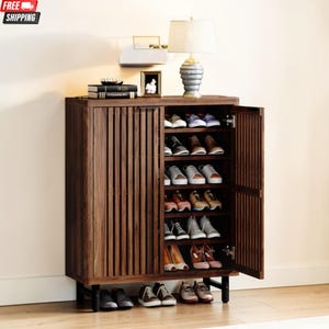 May include: A dark brown wooden shoe cabinet with multiple shelves, holding various pairs of shoes. The cabinet has a vertical slatted design on the doors and sits on black metal legs. A lamp and decorative items are on top.