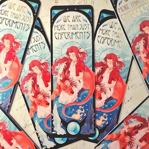 Puede incluir: Marcapáginas artístico con una mujer de pelo rojo y chaqueta roja, sentada sobre una criatura con forma de león. El marcapáginas tiene un borde negro y el texto "WE ARE MORE THAN JUST EXPERIMENTS". La obra de arte incluye detalles en azul, blanco y dorado.