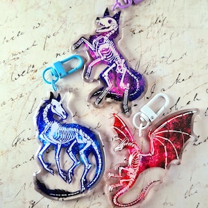 Könnte beinhalten: Drei Schlüsselanhänger aus Acryl mit Skelett-Designs eines Fuchses, eines Einhorns und eines Drachen. Der Fuchs ist lila und rosa, das Einhorn ist blau und der Drache ist rot. Jeder Schlüsselanhänger hat einen Clip.
