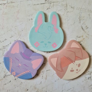 Op de afbeelding: Drie gestanste papieren vormen: een lichtblauw konijn met roze accenten, een paarse kat met een hoed en een perzikkleurige kat. Elk heeft een cartoongezicht. De achtergrond is een vintage papier met vervaagde tekst.