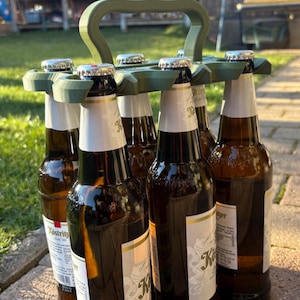 May include: A green plastic beer bottle carrier holding six brown glass beer bottles. The bottles have silver caps and white labels. The carrier has a handle for easy transport. The bottles are 0.5 liters each.