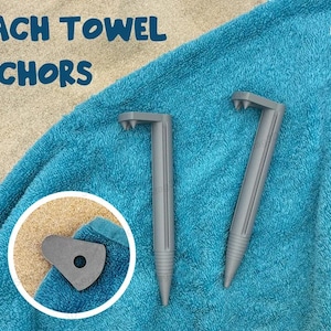 Op de afbeelding: Twee grijze plastic strandhanddoekankers op een blauwe strandhanddoek. De ankers hebben een puntig uiteinde om in het zand te steken en een platte, gebogen bovenkant om de handdoek op zijn plaats te houden. De tekst "BEACH TOWEL ANCHORS" is zichtbaar in de afbeelding.