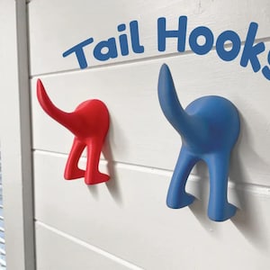 Könnte beinhalten: Zwei schwanzförmige Haken, einer rot und einer blau, an einer weißen Wand montiert. Die Haken sind wie das Hinterteil eines Hundes geformt, mit einem Schwanz und zwei Beinen. Der Text "Tail Hooks" ist in blauen Buchstaben über den Haken geschrieben.
