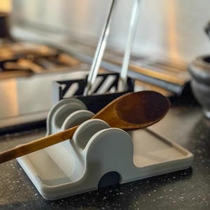 Op de afbeelding: Een grijze siliconen bestekhouder met een houten lepel erop. De houder heeft drie gleuven voor bestek en een platte basis.