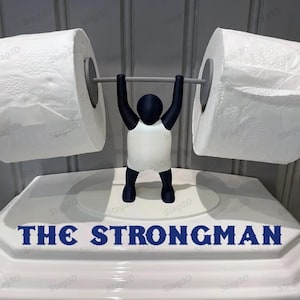 May include: A white toilet paper holder shaped like a person lifting weights. The holder has a blue body and is lifting two rolls of toilet paper. The text "THE STRONGMAN" is printed on the base of the holder.