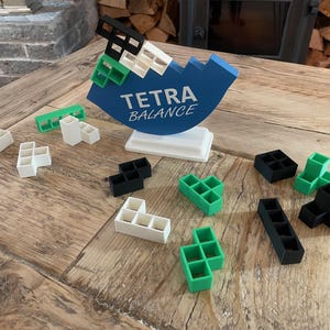 Pode incluir: Um tabuleiro de jogo azul e branco com o texto "TETRA BALANCE" nele. O tabuleiro é cercado por várias peças de plástico pretas, brancas e verdes.