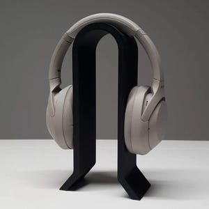 May include: Light grey over-ear headphones displayed on a black headphone stand. The stand has a minimalist design with a curved top and a rectangular base. The headphones are a neutral colour.