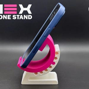 May include: A pink and white phone stand with a blue phone in place. The stand has a curved design with a series of holes along the side. The text "HEX PHONE STAND" is displayed in white and pink.