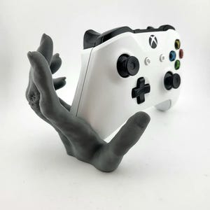 Pode incluir: Uma mão cinza impressa em 3D segurando um controle Xbox branco. O controle tem botões pretos e um logotipo Xbox preto.