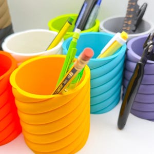 Puede incluir: Un conjunto de seis porta lápices de colores a rayas. Los porta lápices están hechos de un material suave y flexible, perfecto para organizar bolígrafos, lápices y otros artículos de oficina. Los porta lápices están llenos de varios utensilios de escritura, incluidos lápices, bolígrafos y un par de tijeras.