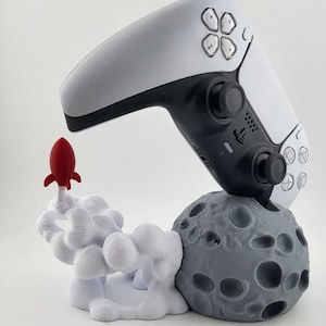 Pode incluir: Um controlador de videogame branco e preto está em um suporte cinza em forma de lua com um foguete vermelho em uma base de nuvem branca.