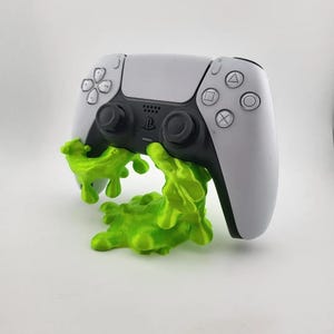 May include: A white PlayStation 5 controller is held in a green 3D printed stand shaped like a blob. The stand has a textured surface.