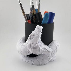 May include: A black pen pot with a white snake-shaped base. The pot holds paintbrushes and art supplies. The snake base has a textured, geometric pattern. The pot is approximately 10 cm in height.