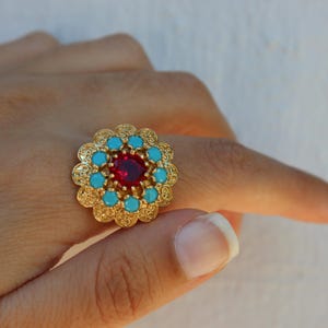 May include: A gold-tone ring featuring a central round red gemstone surrounded by a circular arrangement of small, round, light blue turquoise-colored stones, all set within an intricate, filigree-style scalloped metal frame.