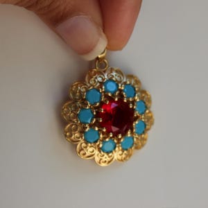 May include: A gold-toned pendant featuring a central faceted red stone surrounded by a ring of small, round, bright blue stones. The metal frame has an intricate, scalloped, filigree-style edge with a circular bail at the top for attachment.