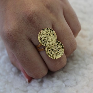 May include: A gold-toned ring featuring two circular coin-shaped charms. Each coin has a textured, sunburst-style border and intricate, embossed markings on the face. The ring band is simple and open, connecting the two overlapping circular elements.