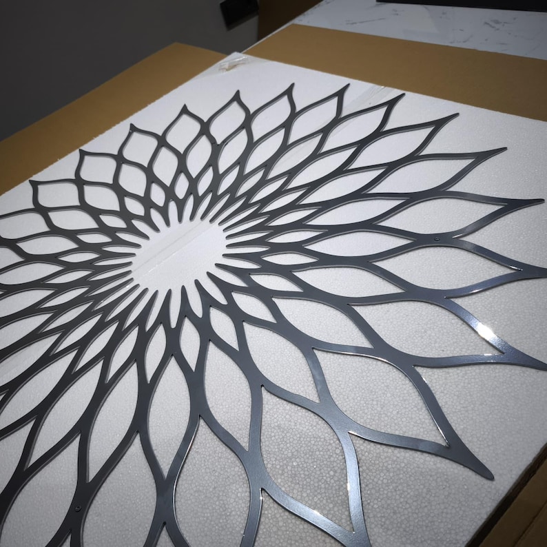 May include: A dark grey metal wall art piece with a floral design. The artwork has a central circular opening and petal-like shapes radiating outwards. It is placed on a white surface.