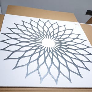 May include: A silver-coloured metal wall art piece with a symmetrical floral design. The artwork is mounted on a white surface. The design is a repeating pattern of petal-like shapes radiating from a central point, creating a flower-like appearance.