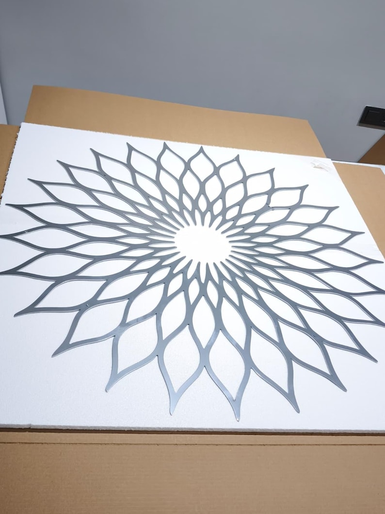 May include: A silver-coloured metal wall art piece with a symmetrical floral design. The artwork is mounted on a white surface. The design is a repeating pattern of petal-like shapes radiating from a central point, creating a flower-like appearance.