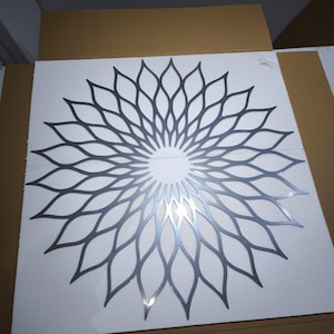 May include: A decorative metal wall art piece in the shape of a stylized flower or sunburst. The design features a series of petal-like shapes radiating from a central circular area. The metal has a silver-grey finish, and the piece is set against a white background.