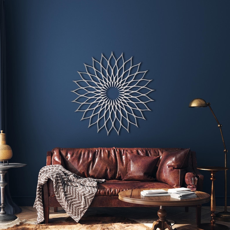 May include: A silver, flower-like wall decoration hangs on a dark blue wall above a brown leather sofa. The room includes a round wooden coffee table and a brass floor lamp. The artwork's design is a series of interconnected petal shapes.
