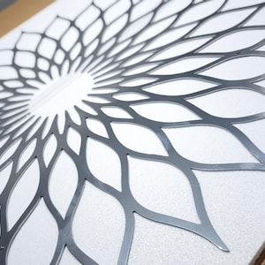 May include: A decorative metal wall art piece with a symmetrical floral design. The artwork is made of silver-coloured metal and has a detailed, openwork pattern of overlapping petal shapes. It is mounted on a white surface.