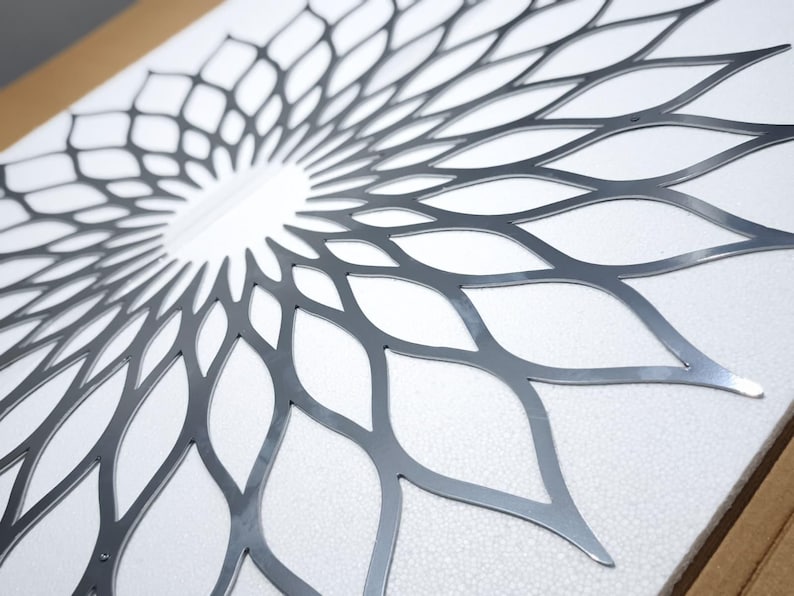 May include: A decorative metal wall art piece with a symmetrical floral design. The artwork is made of silver-coloured metal and has a detailed, openwork pattern of overlapping petal shapes. It is mounted on a white surface.