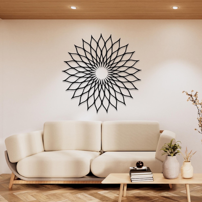 May include: A black, laser-cut metal wall art piece in a floral design, mounted on a white wall above a cream-coloured sofa. The artwork features a circular pattern of petal-like shapes radiating from a central point. The room has a modern, minimalist aesthetic.