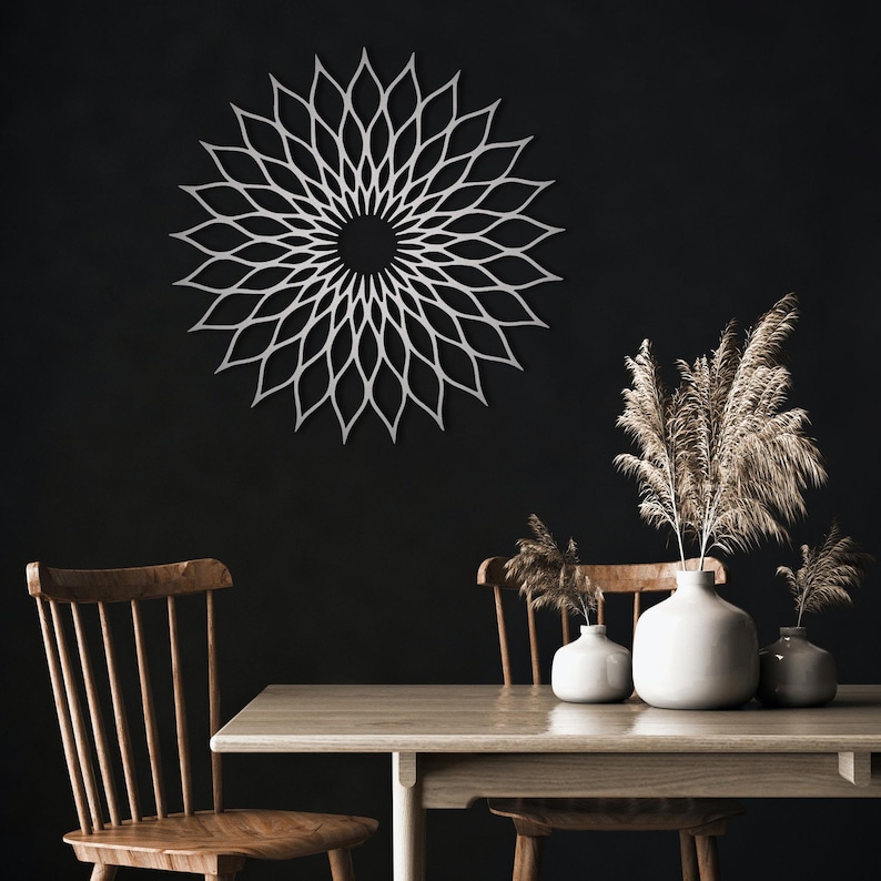 May include: A white metal wall decoration in a floral pattern is mounted on a dark wall. The artwork is the focal point of the room. A wooden table and chairs are in the foreground, with vases of dried plants.