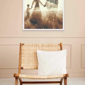 May include: A framed print depicts a family walking through a field, with a man and woman holding hands and a child following. The image has a soft, ethereal quality, with a sepia-toned color palette. A wooden chair with a woven seat and a white pillow is in the foreground.