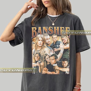 May include: A charcoal gray oversized t-shirt featuring a graphic print of the television show Banshee. The design includes a collage of cast members, some holding firearms, with the word BANSHEE printed in large, bold, orange and yellow letters above.