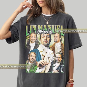 May include: Dark gray t-shirt featuring a graphic of Lin-Manuel Miranda in various poses. The text "Lin Manuel Miranda" is printed in green and white above the images. The shirt is a relaxed fit.